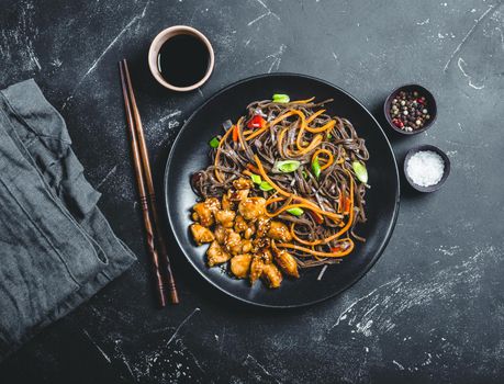 Asian soba noodles, chicken, vegetables, rustic concrete dark background. Soba noodles, teriyaki sauce chicken, vegetables, sesame, chopsticks. Asian style dinner. Chinese/Japanese noodles. Top view