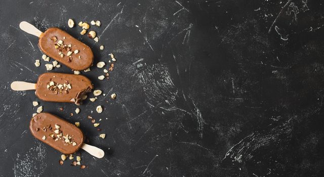 Milk chocolate popsicles with hazelnuts. Close-up. Ice cream popsicles covered with chocolate, sticks, black stone background. Space for text. Top view. Chocolate ice cream bars, nuts. Bitten popsicle