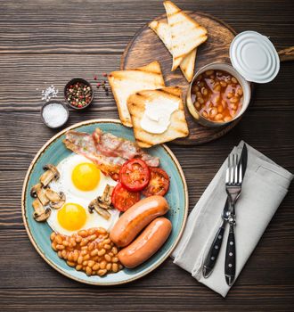 Full English breakfast with fried eggs, sausages, bacon, beans, mushrooms, tomatoes on a plate, bread toasts with butter. Traditional British meal, top view, rustic wooden background.