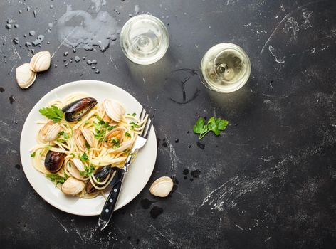 Spaghetti vongole, Italian seafood pasta with clams and mussels, in plate with herbs and two glasses of white wine on rustic stone background, space for text. Italian sea cuisine, close-up, top view