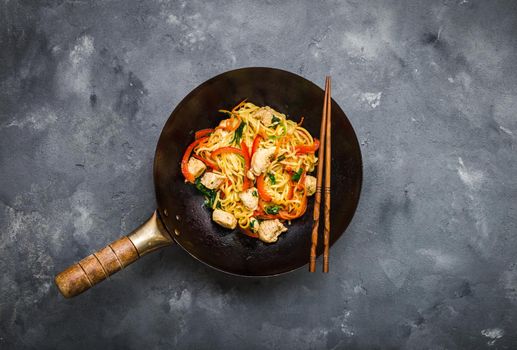 Stir fry noodles, traditional Chinese wok, chopsticks. Asian noodles with vegetables, chicken. Wok noodles, Chinese dinner/lunch. Rustic stone background. Top view. Asian/Chinese noodles. Stir frying.