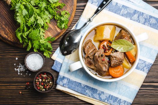 Close-up of slow cooked meat stew ragout in bowl with beef, potato, carrot, broth, herbs, spoon and fork on wooden rustic background, top view. Hot homemade food for dinner, meat casserole .