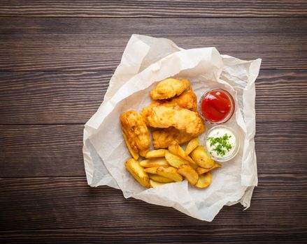 British national traditional fast food fish and chips with assorted dips for choice, on paper, rustic brown wooden background, top view. Battered fried fish, potato chips, tartare and ketchup sauce