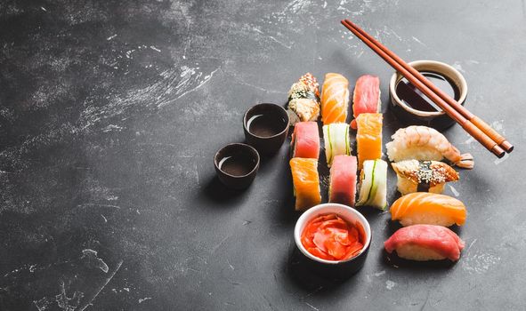 Top view of assorted mixed Japanese sushi set with rolls, nigiri, soy sauce, ginger, chopsticks, two cups of traditional sake on black concrete background. Asian dinner or lunch, free space for text.