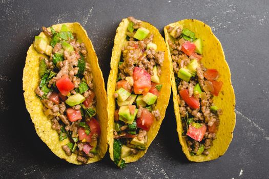Top view, closeup of Mexican tacos with meat, avocado, tomato salsa, herbs in yellow corn tortilla on black rustic stone background. Traditional Mexican dish for lunch or dinner, concept