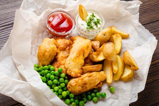 Close-up of British traditional fast food fish and chips with assorted dips, peas, on paper, rustic brown wooden background. Battered fried fish, potato chips, tartare and ketchup sauce