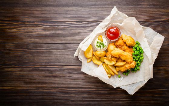 British traditional fast food fish and chips with dips for choice, peas, on paper, rustic wooden background, top view, space for text. Battered fried fish, potato chips, tartare and ketchup sauce