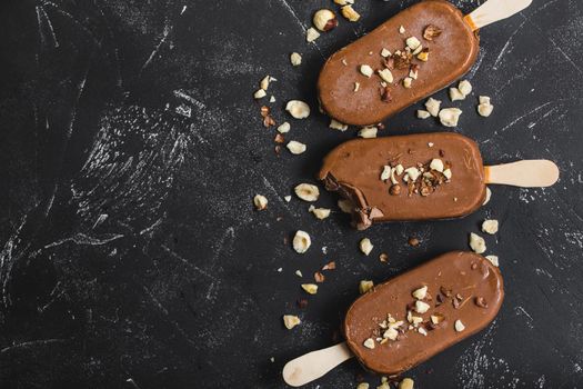 Milk chocolate popsicles with hazelnuts. Close-up. Ice cream popsicles covered with chocolate, sticks, black stone background. Space for text. Top view. Chocolate ice cream bars, nuts. Bitten popsicle