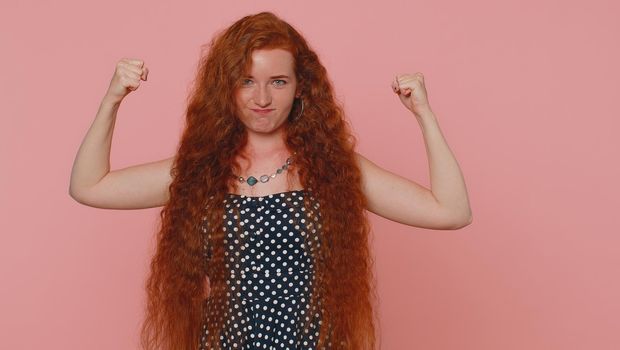I am strong and independent. Young redhead woman showing biceps and looking confident, feeling power strength to fight for rights, energy to gain success win. Ginger girl on pink studio background