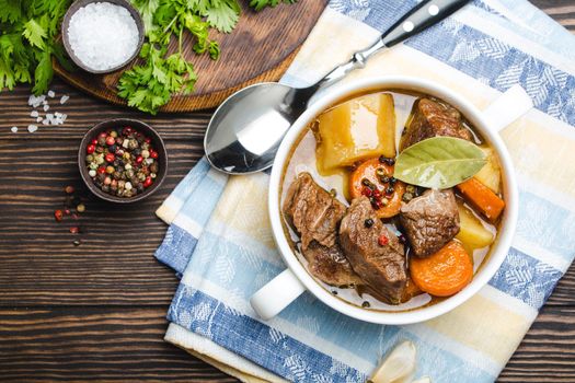 Close-up of slow cooked meat stew ragout in bowl with beef, potato, carrot, broth, herbs and spoon on wooden rustic background, top view. Hot homemade food for dinner, meat casserole .