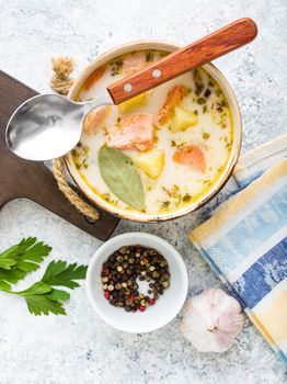 Salmon soup with potatoes, carrot, cream, herbs. Scandinavian/Norwegian fish soup in bowl, cutting wooden board, spoon, rustic white concrete background. Salmon soup for dinner. Top view. Close-up