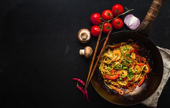 Stir fry noodles in traditional Chinese wok, chopsticks, ingredients. Space for text. Asian noodles with vegetables, shrimps. Wok noodles. Black dark background. Top view. Asian/Chinese dinner