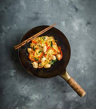 Stir fry noodles, traditional Chinese wok, chopsticks. Asian noodles with vegetables, chicken. Wok noodles, Chinese dinner/lunch. Rustic stone background. Top view. Asian/Chinese noodles. Stir frying.