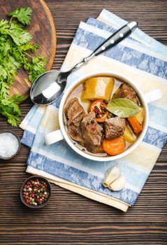 Close-up of slow cooked meat stew ragout in bowl with beef, potato, carrot, broth, herbs and spoon on wooden rustic background, top view. Hot homemade food for dinner, meat casserole .