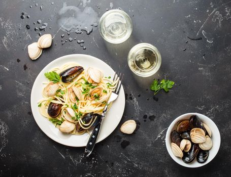 Spaghetti vongole, Italian seafood pasta with clams and mussels, in plate with herbs and two glasses of white wine on rustic stone background. Traditional Italian sea cuisine, close-up, top view