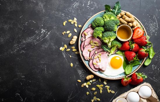 Ketogenic low carbs diet, top view, space for text. Plate on stone black background with keto foods: egg, meat, olive oil, broccoli, berries, nuts, seeds. Healthy fats, clean eating for weight loss