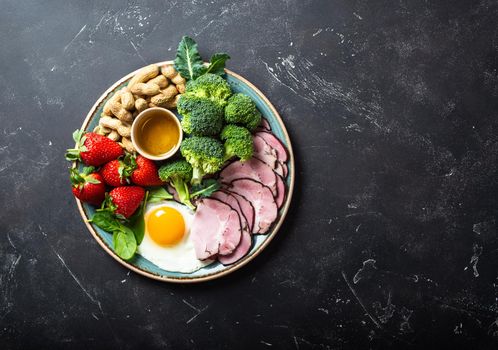 Ketogenic low carbs diet, top view, space for text. Plate on stone black background with keto foods: egg, meat, olive oil, broccoli, berries, nuts, seeds. Healthy fats, clean eating for weight loss