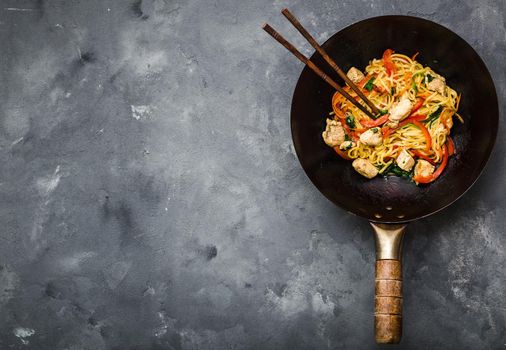 Stir fry noodles in traditional Chinese wok, chopsticks, ingredients. Space for text. Asian noodles with vegetables, chicken. Wok noodles. Rustic stone background. Top view. Asian/Chinese dinner