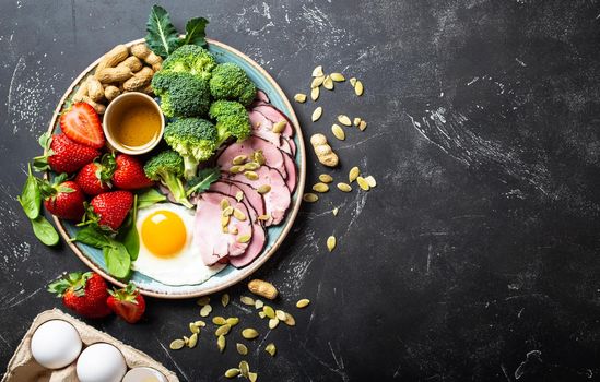 Ketogenic low carbs diet, top view, space for text. Plate on stone black background with keto foods: egg, meat, olive oil, broccoli, berries, nuts, seeds. Healthy fats, clean eating for weight loss