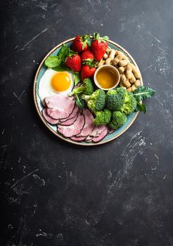 Ketogenic low carbs diet, top view, space for text. Plate on stone black background with keto foods: egg, meat, olive oil, broccoli, berries, nuts, seeds. Healthy fats, clean eating for weight loss