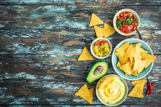 Mexican tortilla chips with guacamole, cheese, salsa dips. Space for text. Mexican party food, appetizers. Top view. Tortilla chips, assorted sauces, hot pepper, avocado, rustic wooden background