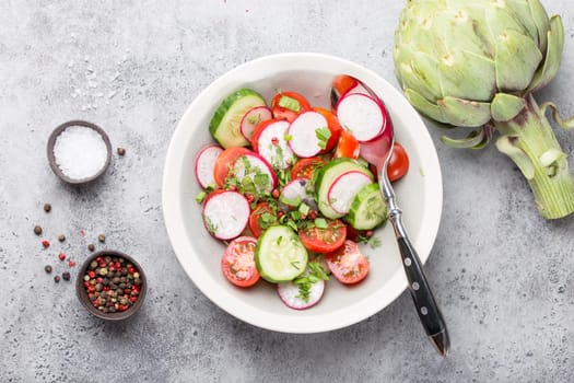 Close-up of fresh healthy salad in a bowl made of tomatoes, cucumber, radish and herbs, with raw artichoke and seasonings, good for diet or detox, grey rustic stone background, top view