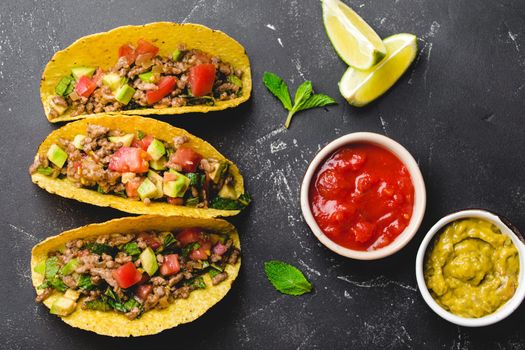 Top view of Mexican tacos with meat, vegetables in yellow corn tortilla served with lime, salsa and guacamole sauces on black rustic stone background. Traditional Mexican dish for lunch or dinner