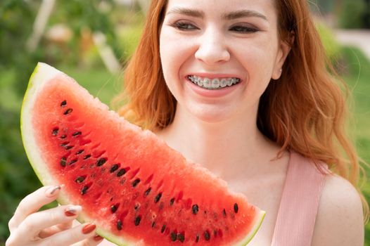 Beautiful red-haired woman smiling with braces and about to eat a slice of watermelon outdoors in summer.