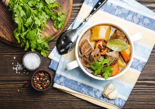 Close-up of slow cooked meat stew ragout in bowl with beef, potato, carrot, broth, herbs and spoon on wooden rustic background, top view. Hot homemade food for dinner, meat casserole .