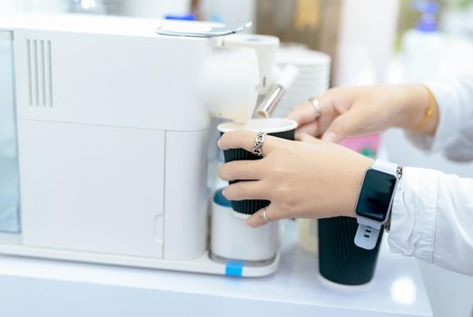 Woman making a cup of hot coffee with capsule coffee machine. Woman holding black cup taking coffee from a capsule coffee machine on the table. Espresso maker. Morning drink. Modern home equipment.