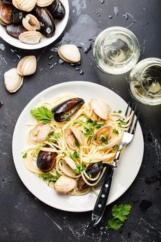 Spaghetti vongole, Italian seafood pasta with clams and mussels, in plate with herbs and two glasses of white wine on rustic stone background. Traditional Italian sea cuisine, close-up, top view