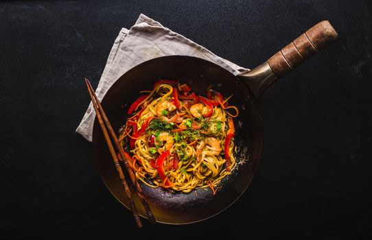 Stir fry noodles in traditional Chinese wok, chopsticks. Asian noodles with vegetables, shrimps. Wok noodles, Chinese dinner/lunch. Black dark background. Top view. Asian/Chinese noodles. Stir frying