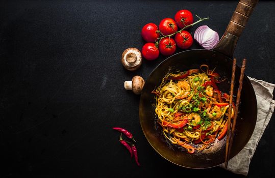 Stir fry noodles in traditional Chinese wok, chopsticks, ingredients. Space for text. Asian noodles with vegetables, shrimps. Wok noodles. Black dark background. Top view. Asian/Chinese dinner