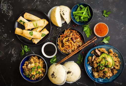 Asian assorted food set, dark rustic stone background. Chinese dishes. Chinese stir-fry noodles, asian rice with meat, dim sum, fried spring rolls, steamed Chinese buns. Top view. Asian stile dinner