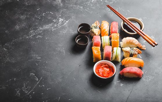Top view of assorted mixed Japanese sushi set with rolls, nigiri, soy sauce, ginger, chopsticks, two cups of traditional sake on black concrete background. Asian dinner or lunch, free space for text.