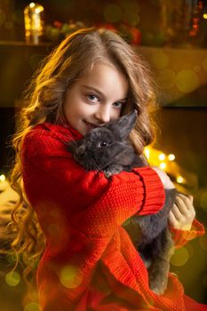 A little blonde girl with a gray rabbit in her arms next to a Christmas tree decorated with garlands, glass balls and Christmas toys and a fireplace. The concept of winter holidays is Christmas and New Year holidays. Magical festive atmosphere.