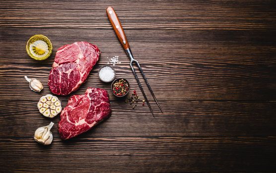 Raw marbled meat steak Ribeye on rustic wooden background with fork, seasonings and olive oil for cooking, roasting, grill or BBQ party with space for text, top view. Meat restaurant concept