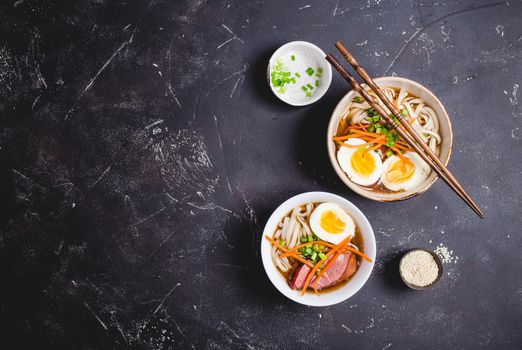 Bowls with Japanese soup ramen, chopsticks, black concrete rustic background. Traditional Asian soup with noodles, meat, broth, eggs. Asian food. Japanese ramen for dinner. Ramen, close-up. Overhead.