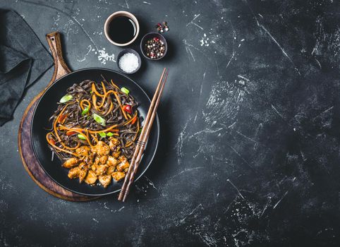Asian soba noodles, chicken, vegetables, dark background. Space for text. Soba noodles, teriyaki sauce chicken, vegetables, sesame, chopsticks. Asian style dinner. Chinese/Japanese noodles. Top view