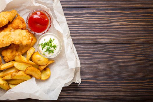 British traditional fast food fish and chips with assorted dips for choice, on paper, rustic wooden background, top view, space for text. Battered fried fish, potato chips, tartare and ketchup sauce