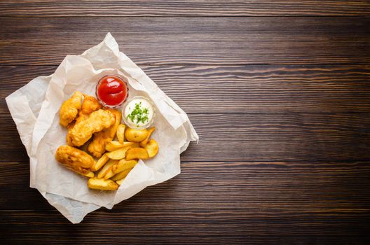 British traditional fast food fish and chips with assorted dips for choice, on paper, rustic wooden background, top view, space for text. Battered fried fish, potato chips, tartare and ketchup sauce