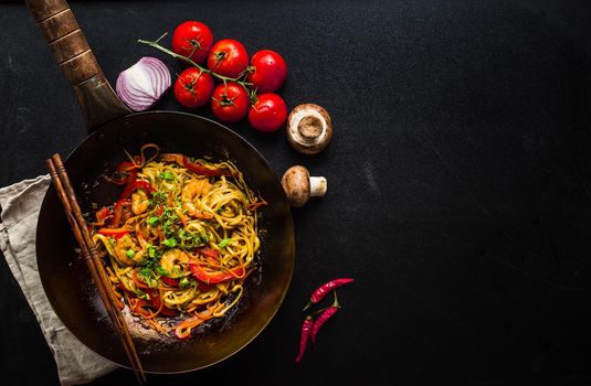 Stir fry noodles in traditional Chinese wok, chopsticks, ingredients. Space for text. Asian noodles with vegetables, shrimps. Wok noodles. Black dark background. Top view. Asian/Chinese dinner