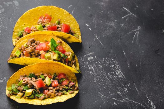 Top view of Mexican tacos with meat, tomato salsa, avocado, herbs in yellow corn tortilla on black rustic stone background with copy space. Traditional Mexican dish for lunch or dinner