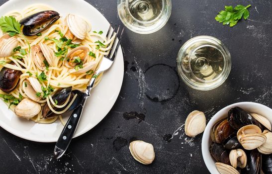 Spaghetti vongole, Italian seafood pasta with clams and mussels, in plate with herbs and two glasses of white wine on rustic stone background. Traditional Italian sea cuisine, close-up, top view