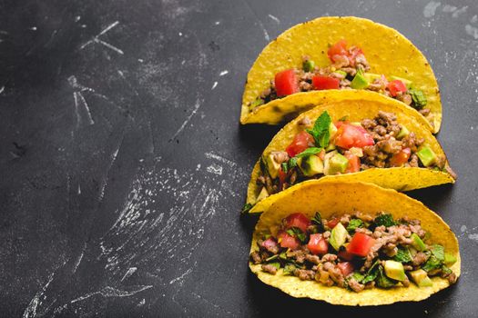 Top view of Mexican tacos with meat, tomato salsa, avocado, herbs in yellow corn tortilla on black rustic stone background with copy space. Traditional Mexican dish for lunch or dinner