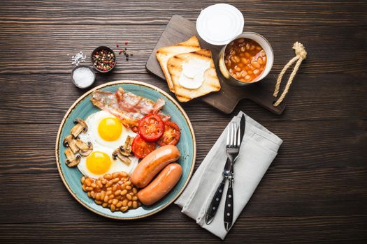 Full English breakfast with fried eggs, sausages, bacon, beans, mushrooms, tomatoes on a plate, bread toasts with butter. Traditional British meal, top view, rustic wooden background.