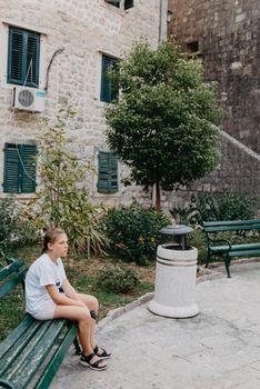 Girl Tourist Resting in the Ancient Narrow Street On A Beautiful Summer Day In MEDITERRANEAN MEDIEVAL CITY, OLD TOWN KOTOR, MONTENEGRO. Young Beautiful Cheerful Woman Walking On Old Street