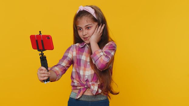 Wow reaction. Impressed young preteen child girl kid blogger taking selfie on mobile phone selfie stick, communicating video call online. Little toddler children isolated on studio yellow background