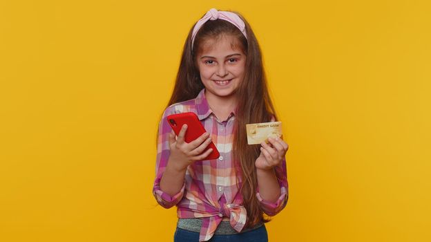Young preteen child girl kid using credit bank card and smartphone while transferring money, purchases online shopping, order food delivery. Little toddler children alone on studio yellow background