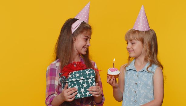 Happy little toddler children sister girls siblings friends wearing festive cone cap celebrating birthday anniversary party, blowing candle on small cake cupcake making a wish, dancing. Preschool kids
